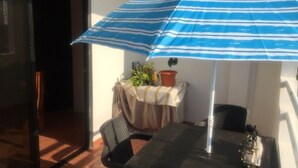 Uma área de pátio com mesa e cadeiras, um guarda-sol listrado de azul e um vaso com planta.