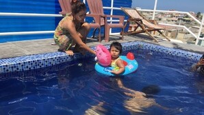 Uma área de piscina com uma criança boiando, uma mulher e cadeiras de descanso.