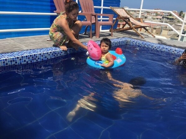 Une zone de piscine avec un enfant sur un flotteur, une femme et des chaises longues.