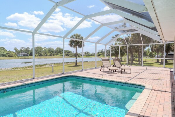 A pool area with a clear view of a lake, surrounded by a covered deck with lounge chairs. - Maria Maria | Lake View Home with Heated Pool (Lehigh Acres)