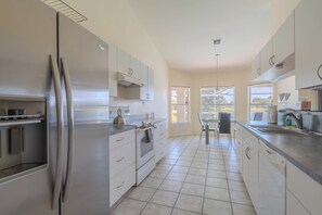 A modern kitchen with stainless steel appliances, a dining area with a table and chairs, and large windows providing natural light. - Maria Maria | Lake View Home with Heated Pool (Lehigh Acres)