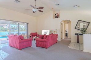 A spacious living room with a large window overlooking a pool, two pink sofas, a ceiling fan, and a view of the pool area. - Maria Maria | Lake View Home with Heated Pool (Lehigh Acres)