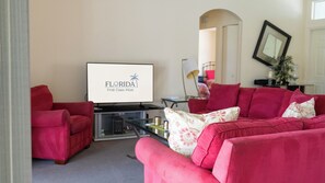 Un saló amb una televisió de pantalla plana que mostra 'FLORIDA First Class Villas', un sofà vermell, una taula de centre i una làmpada.