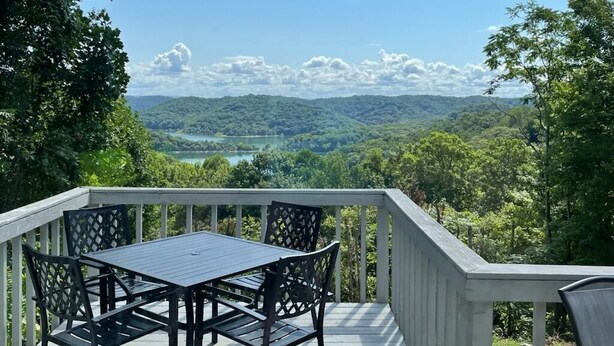 A deck with a table and chairs overlooking a lake and forested hills.