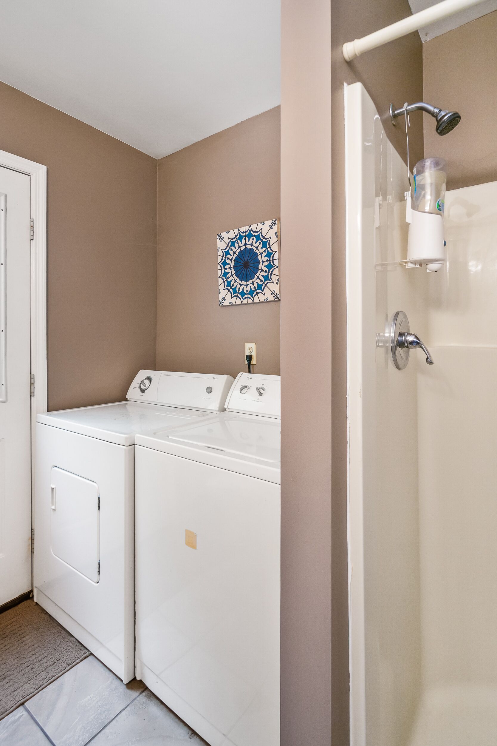 Zona de lavandería con dos lavadoras blancas, ducha con puerta de vidrio y un azulejo decorativo en la pared.