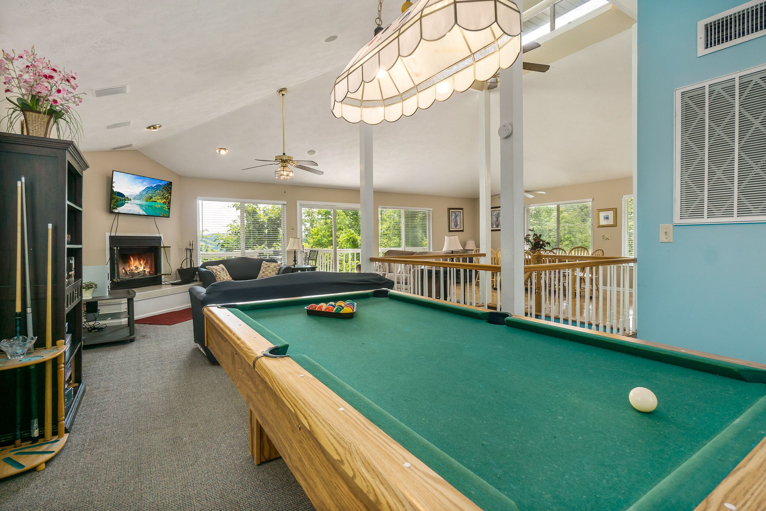 A pool table with a ball, a sofa, a fireplace, and a TV in a well-lit room with large windows.