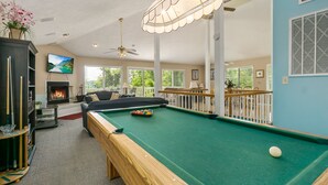 A pool table with a ball, a sofa, a fireplace, and a TV in a well-lit room with large windows.