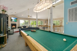 Une table de billard avec des billes, un canapé, une cheminée et un téléviseur dans une pièce bien éclairée, dotée de grandes fenêtres.