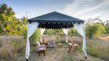 A tent with a canopy and white drapes, surrounded by outdoor furniture including two chairs and a table, set in a grassy area with trees in the background.