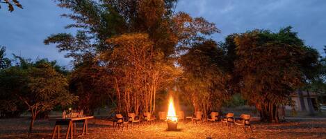 An outdoor seating area with a fire pit surrounded by chairs, set in a natural landscape with trees.
