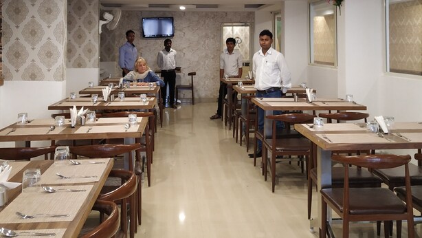 A restaurant with multiple tables and chairs, a flat-screen TV mounted on the wall, and a ceiling with recessed lighting. - Aum Hotel (Puttaparti)