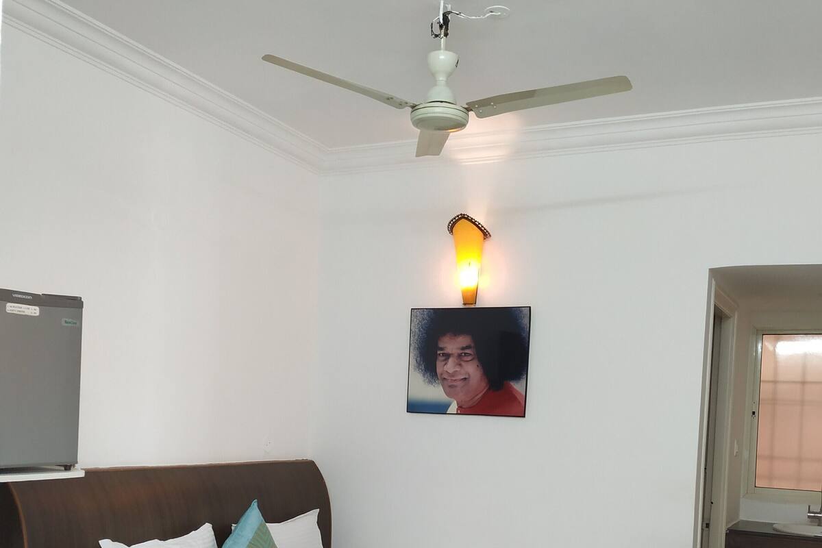 Une chambre à coucher avec un lit, un ventilateur de plafond, une lampe fixée au mur, un tableau et un téléviseur posé sur un meuble.