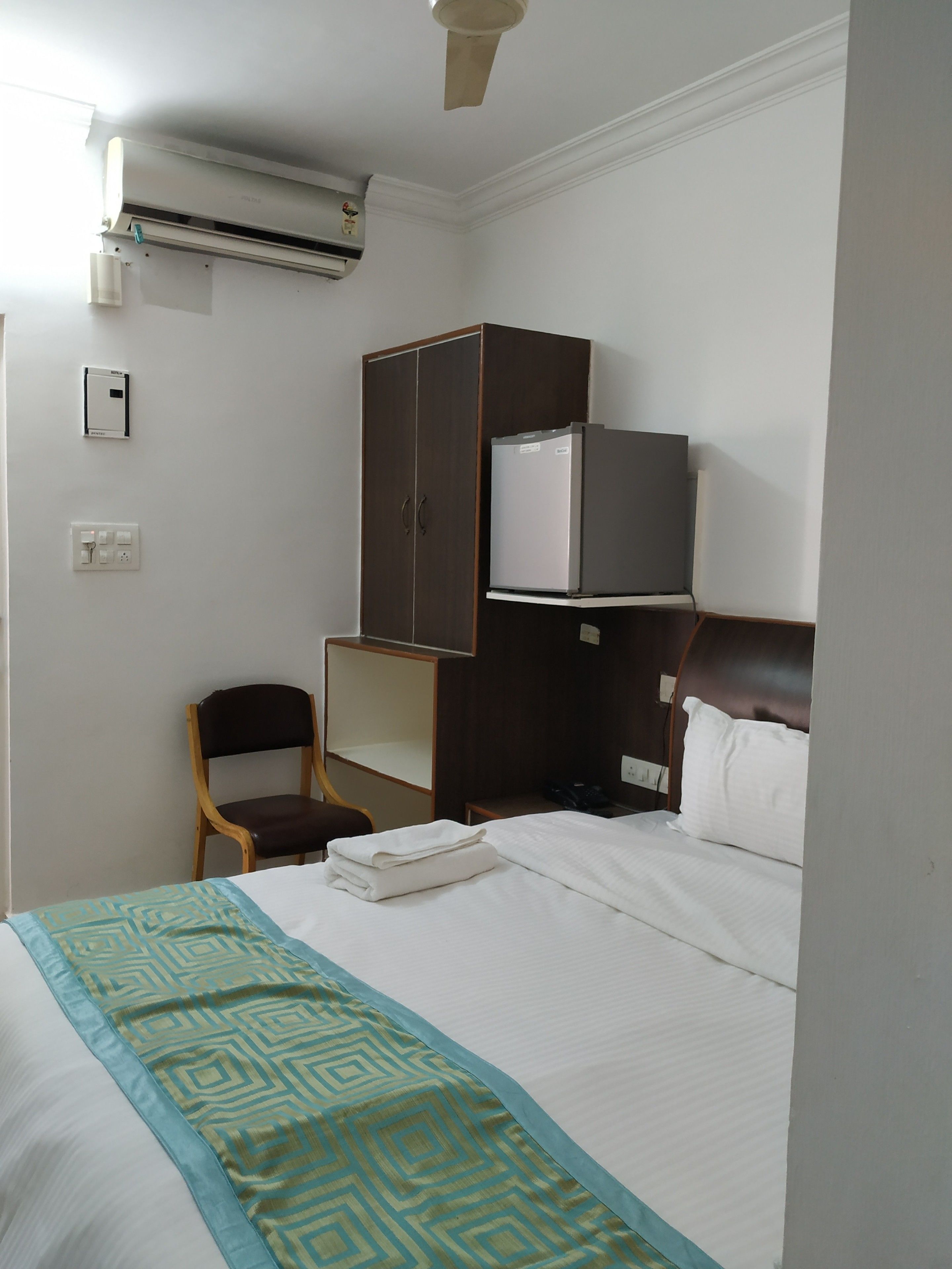 A hotel room with a bed, a wardrobe, a chair, a desk, an air conditioner, and a ceiling fan.
