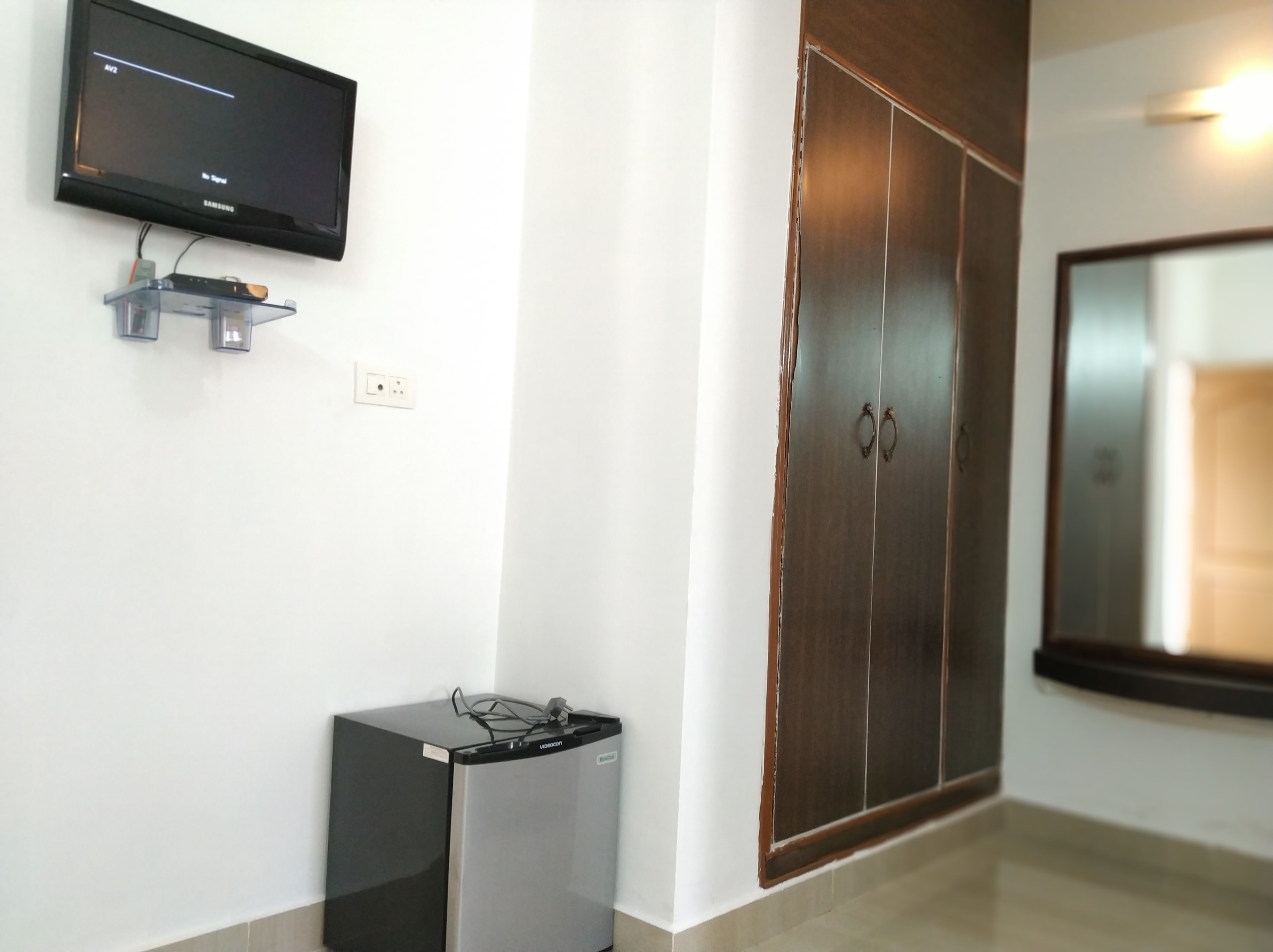 A room with a television mounted on the wall, a small refrigerator, and a wardrobe with a mirror.