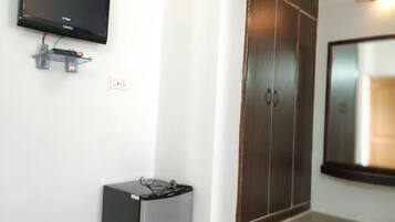 A room with a television mounted on the wall, a small refrigerator, and a wardrobe with a mirror.