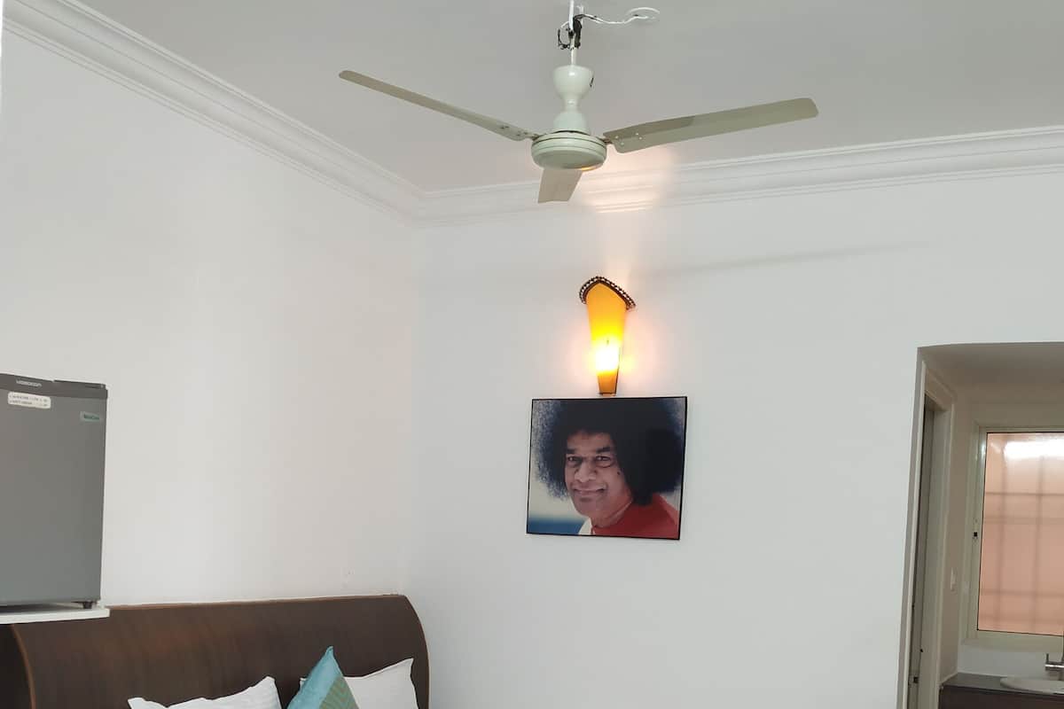 Un lit bien fait, avec du linge de lit blanc et un couvre-lit à motifs, un ventilateur de plafond, une lampe fixée au mur et un tableau encadré au-dessus du lit.