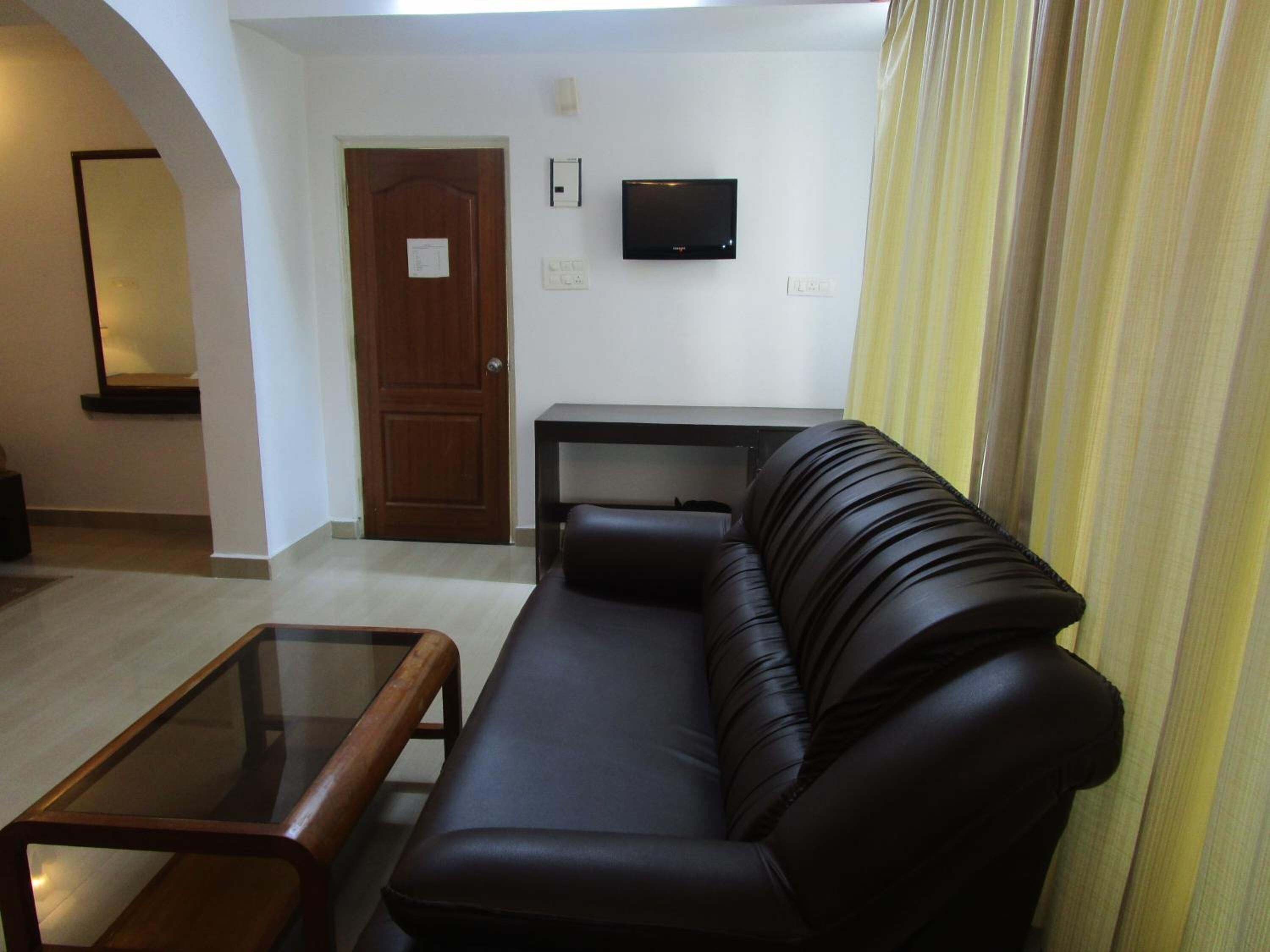 A living room with a black leather sofa, a glass coffee table, a wooden door, a flat-screen TV mounted on the wall, and a curtain on the right.