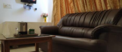A living room with a brown leather sofa, a wooden coffee table, a television mounted on the wall, and a window with curtains.