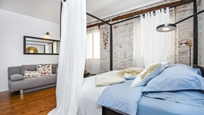 A bedroom with a bed, a grey sofa, a white curtain, a mirror, and a stone wall. - Sunrise Sea view (Rovinj)