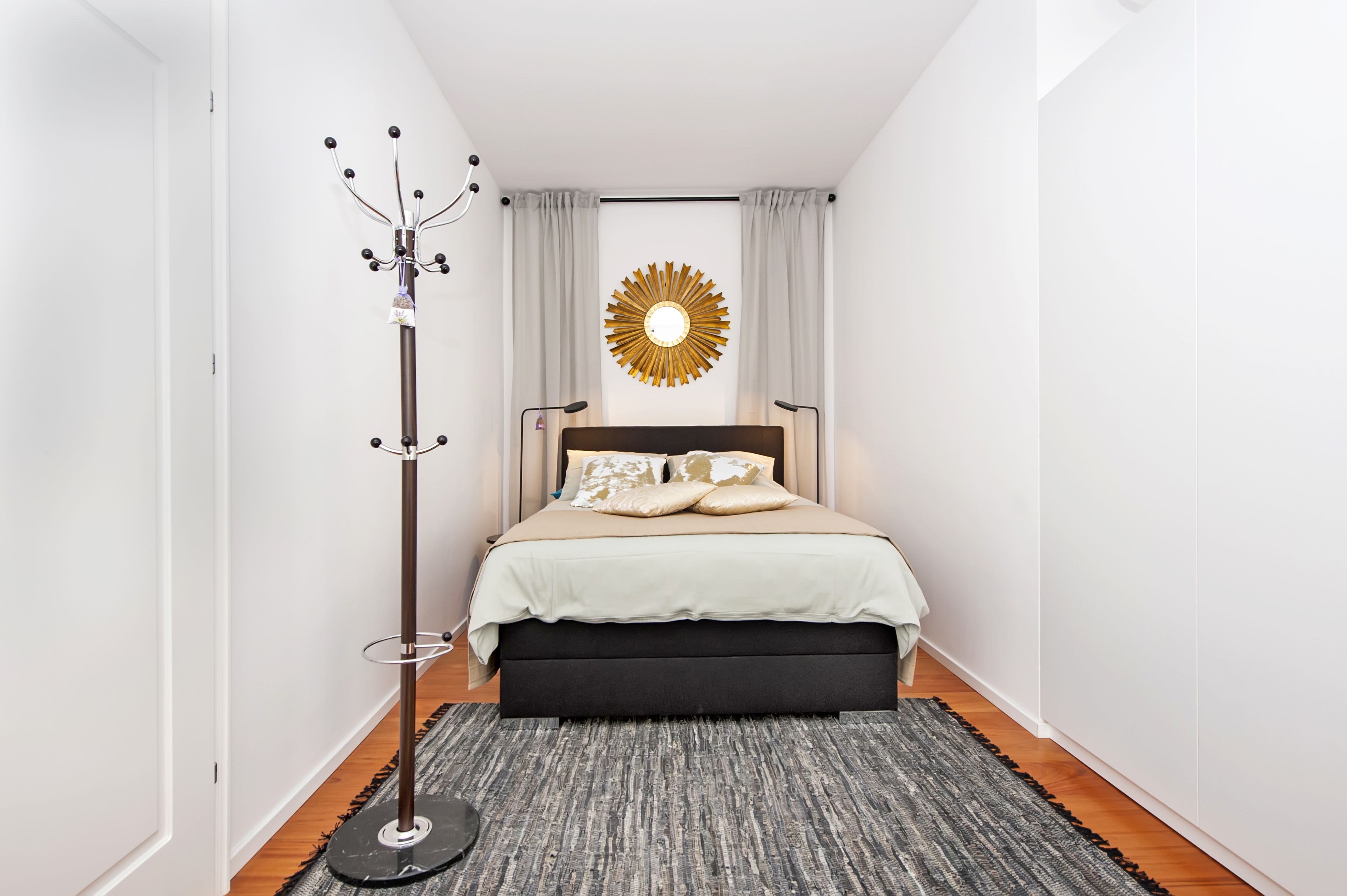A bedroom with a bed, a coat rack, a wall-mounted sunburst decoration, and a grey rug.