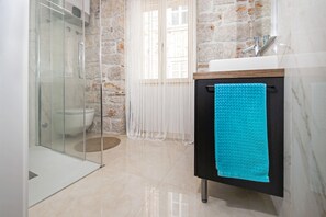 Une salle de bain avec une douche à parois de verre, un mur en pierre, un rideau blanc, un lavabo avec une serviette bleue et un sol en marbre.
