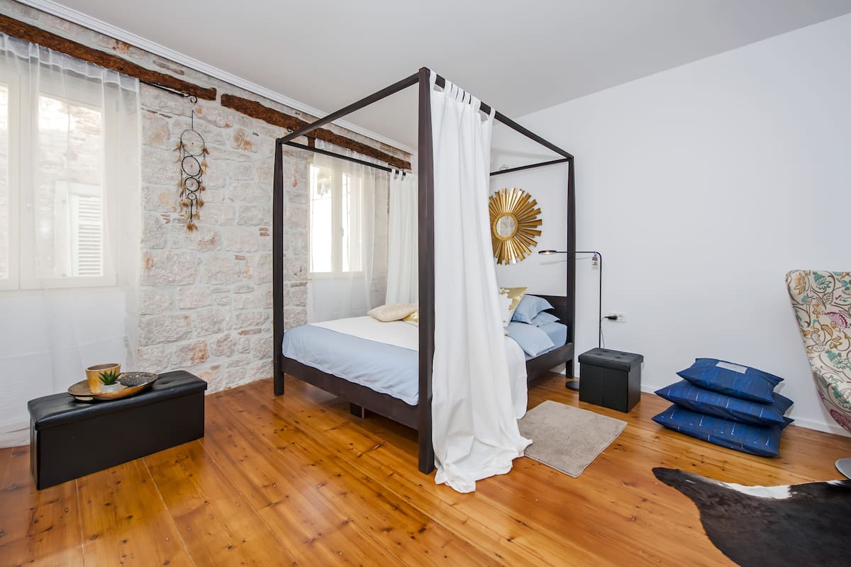 A bedroom with a four-poster bed, stone wall, wooden flooring, and a small bedside table with a lamp.