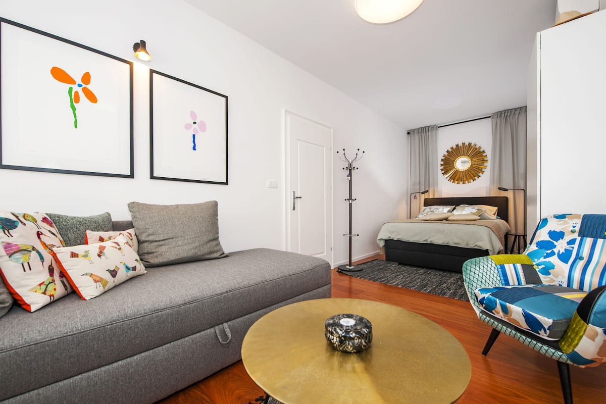 A modern living room with a grey sofa, colorful pillows, a round coffee table, a bed with a headboard, and a decorative wall piece.