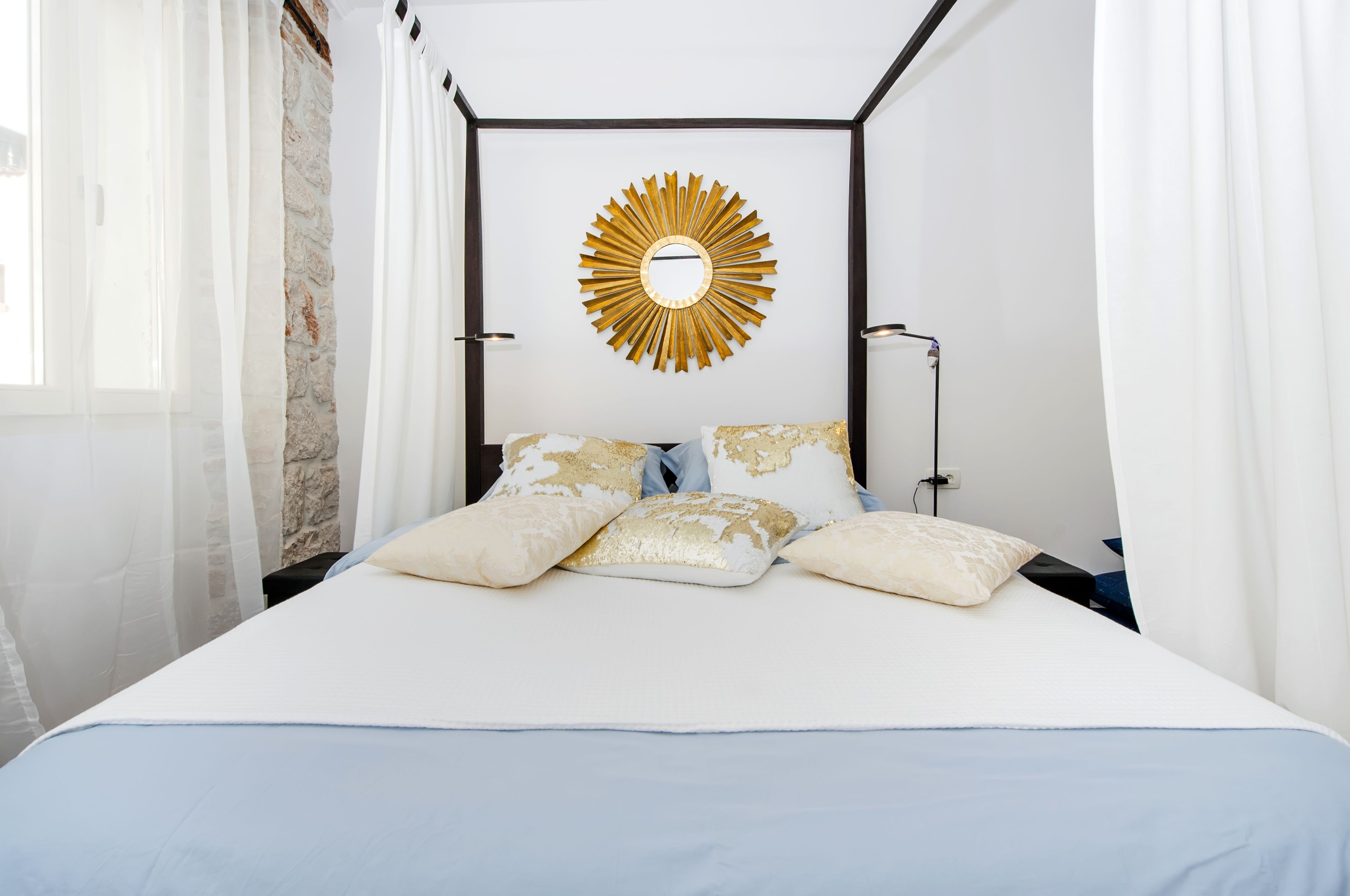 Een net opgemaakt bed met wit-en-gouden beddengoed, een zonneschijf spiegel erboven en een stenen muur achter.