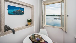 A room with a view of a harbor, a framed painting, a small plant, and a table with decorative items. - Sunrise Sea view (Rovinj)