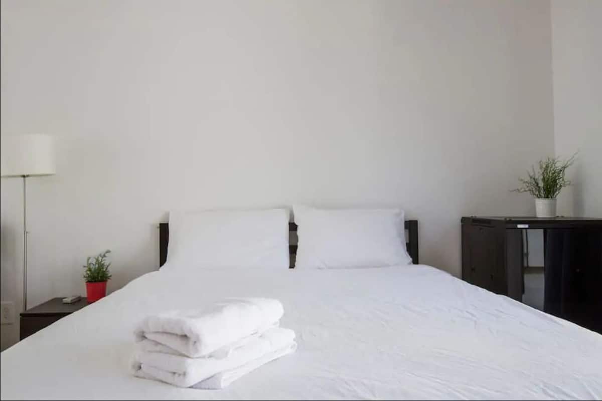 A neatly made bed with white linens and a stack of folded towels. There is a small potted plant on a nightstand beside the bed.