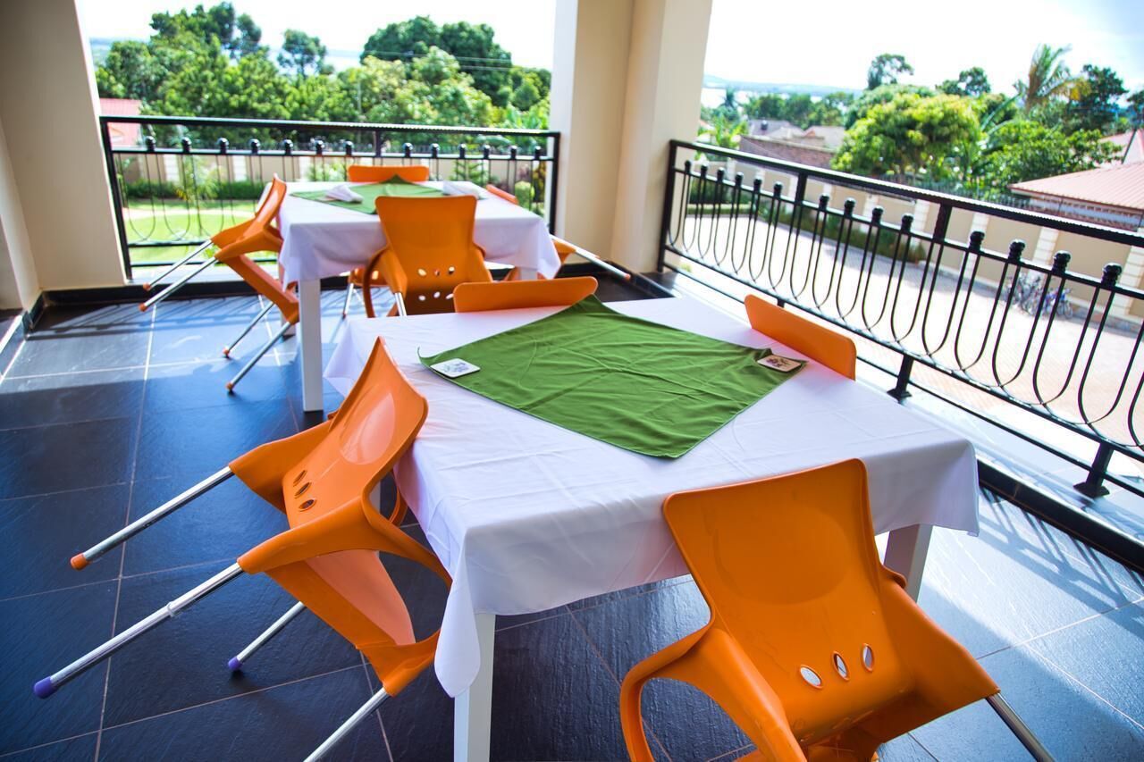 A balcony with tables and orange chairs, a green mat, and a view of greenery and buildings.