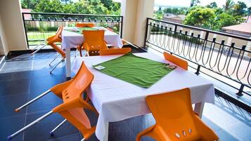 A balcony with tables and orange chairs, a green mat, and a view of greenery and buildings.