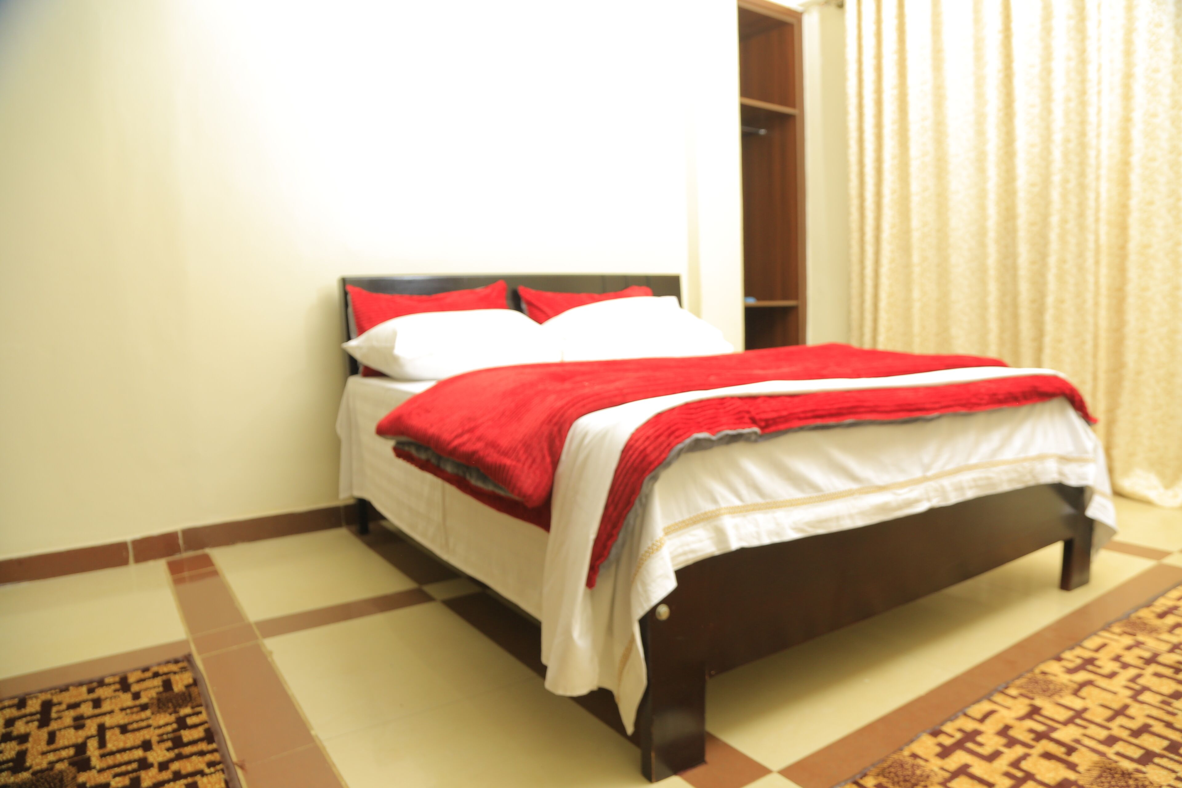 A neatly made bed with a red and white bedspread, a wooden headboard, and a patterned rug on a tiled floor.