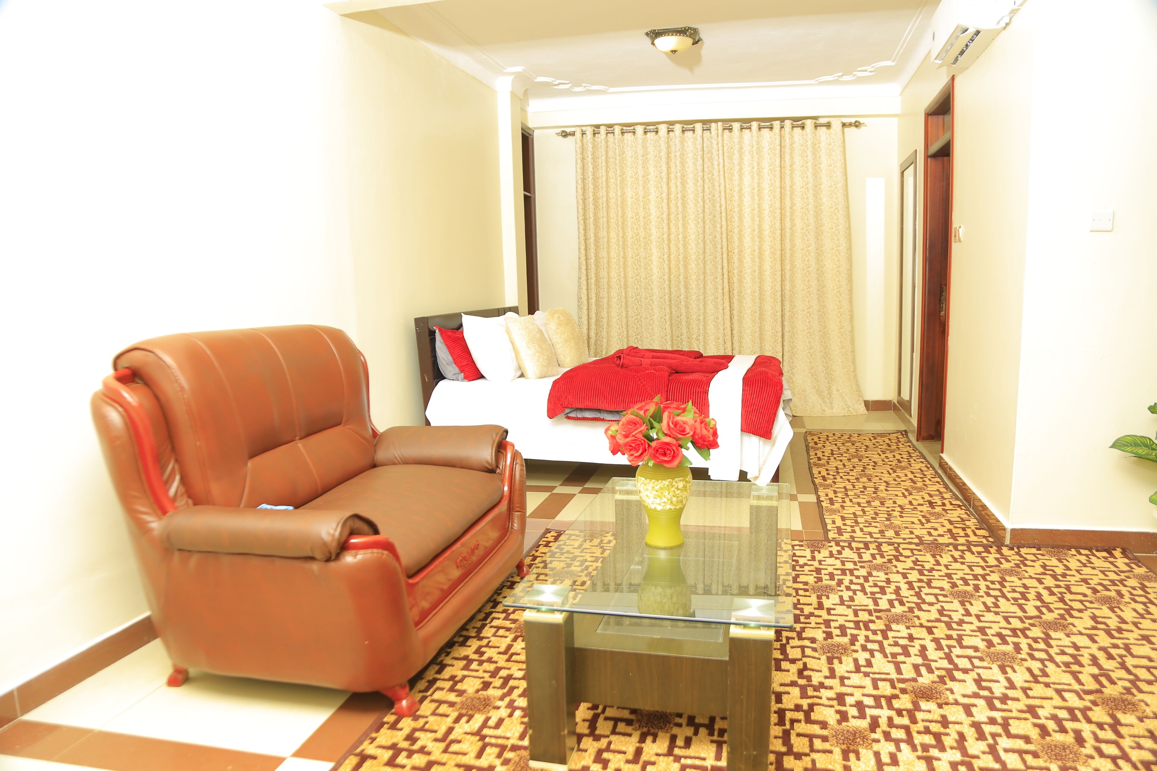A hotel room with a brown leather sofa, a glass coffee table with a vase of flowers, a bed with a red blanket, and a patterned carpet.