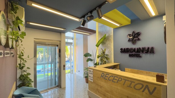 Ein Hotel-Empfangsbereich mit einem Holzbüro, einem Schild mit der Aufschrift "SARDUMYA HOTEL" und einer Empfangstheke. Um den Empfangsbereich befinden sich Stühle und Kübelpflanzen.