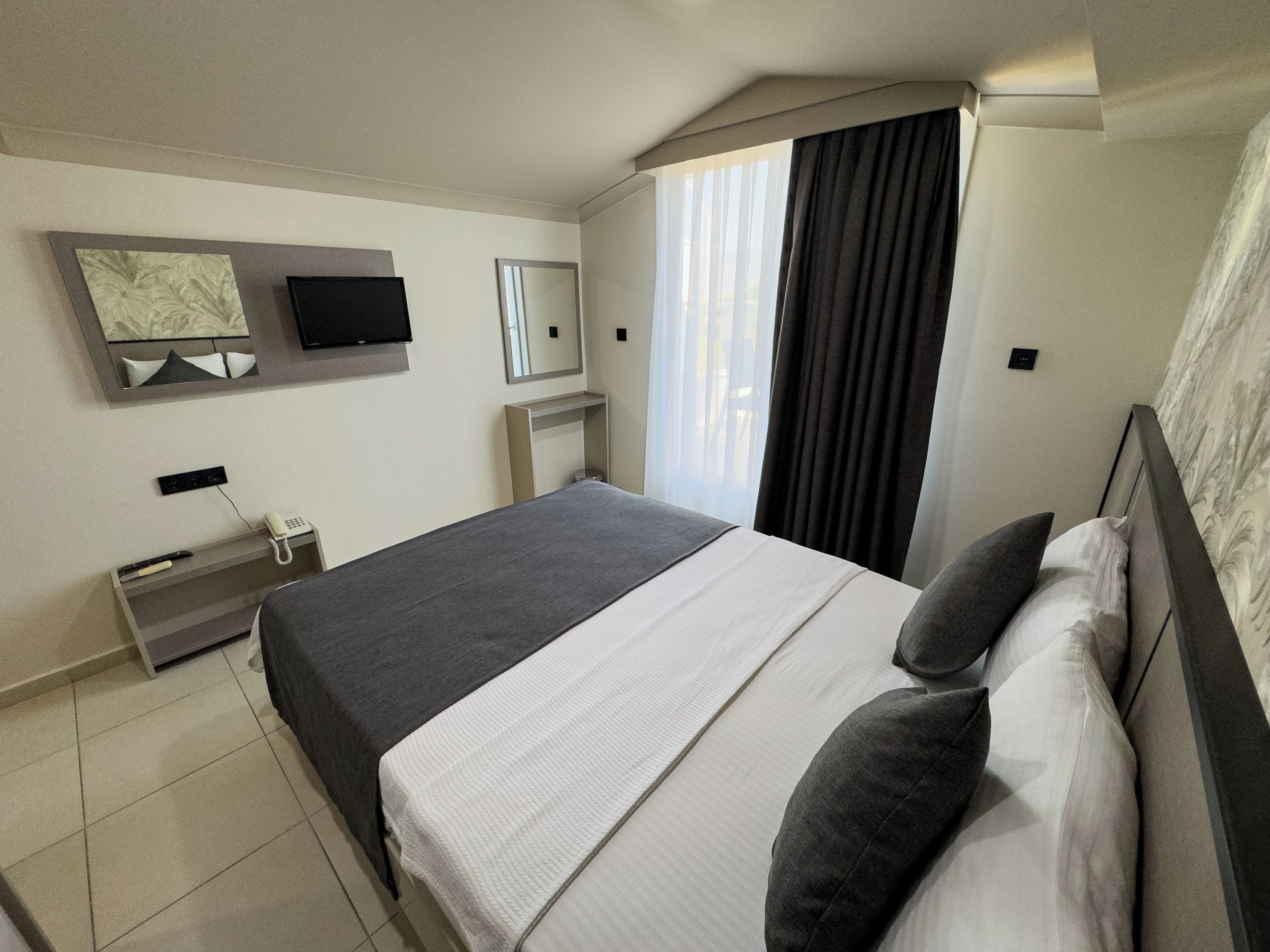 A modern hotel room with a large bed, a flat-screen TV mounted on the wall, a bedside table with a phone, and a window with curtains.