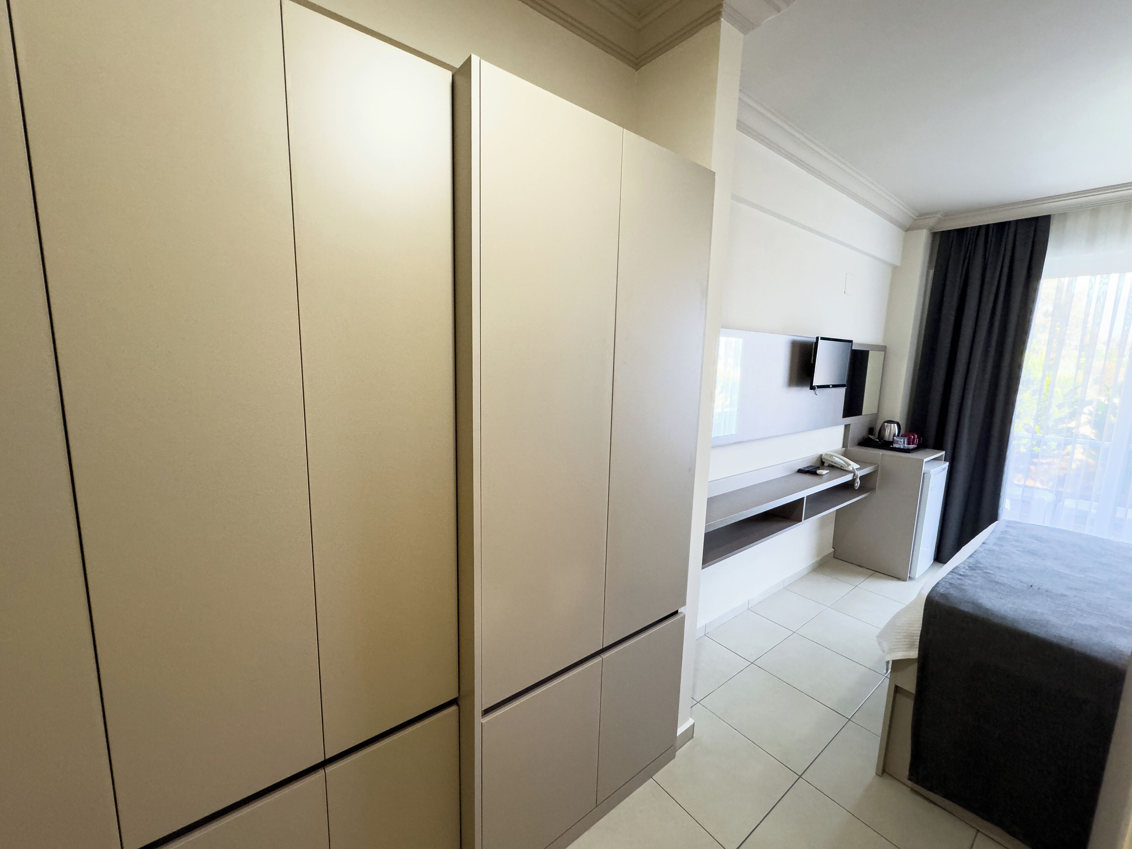 A hotel room with a large wardrobe, a flat-screen TV mounted on the wall, a small desk with a telephone, and a bed with a dark cover.