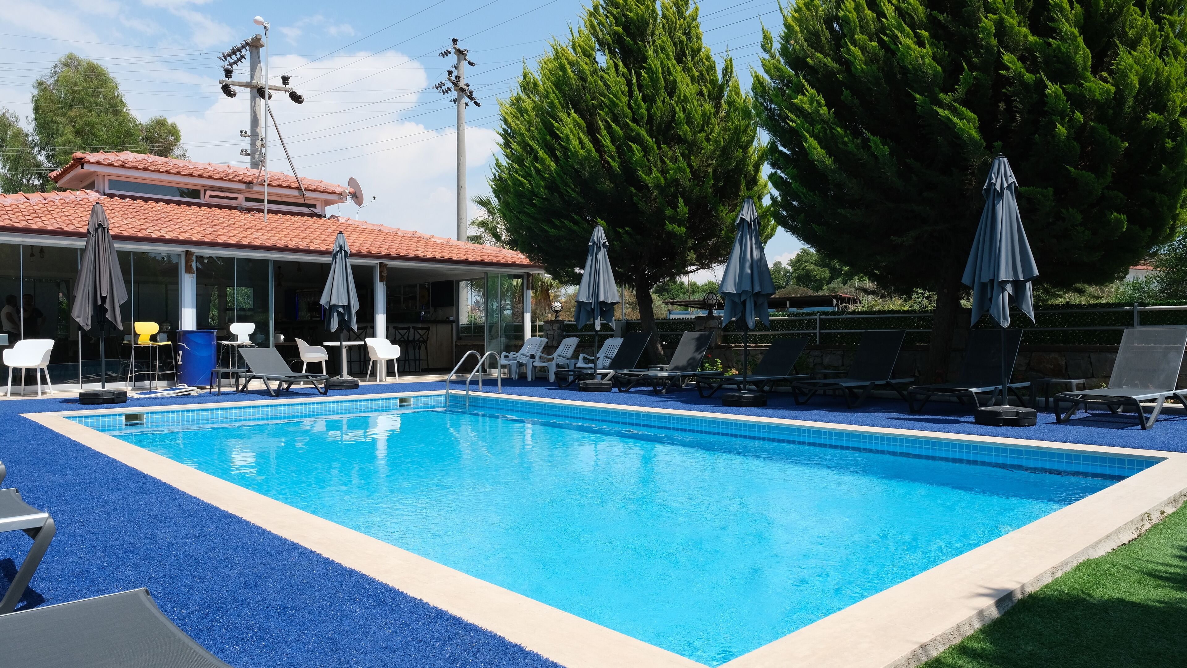 A rectangular swimming pool with blue tiles, surrounded by lounge chairs and umbrellas, and a building with a tiled roof in the background.