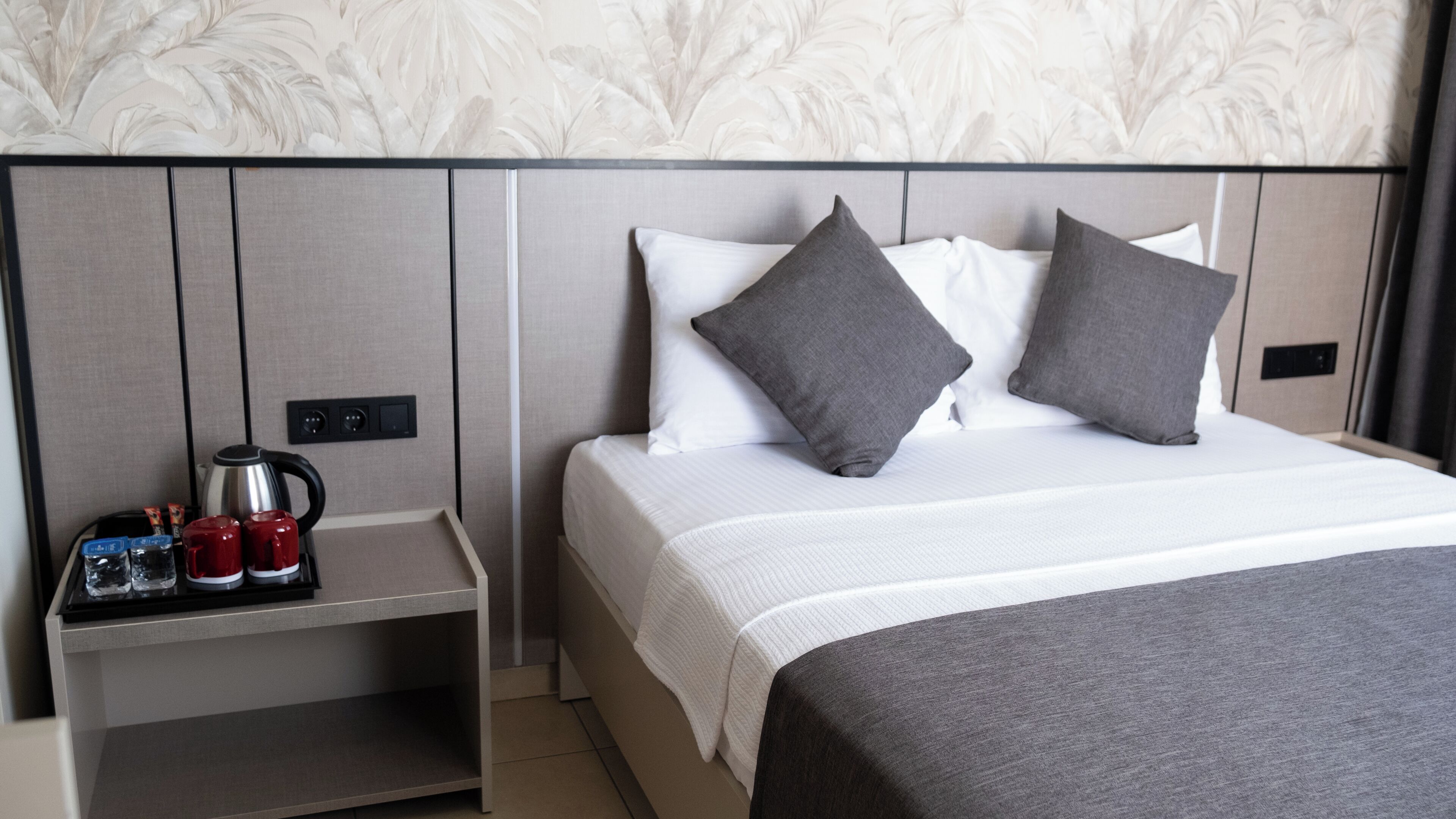 A neatly made bed with grey and white bedding, a bedside table with a kettle, cups, and a tray, and a wall with a floral pattern.