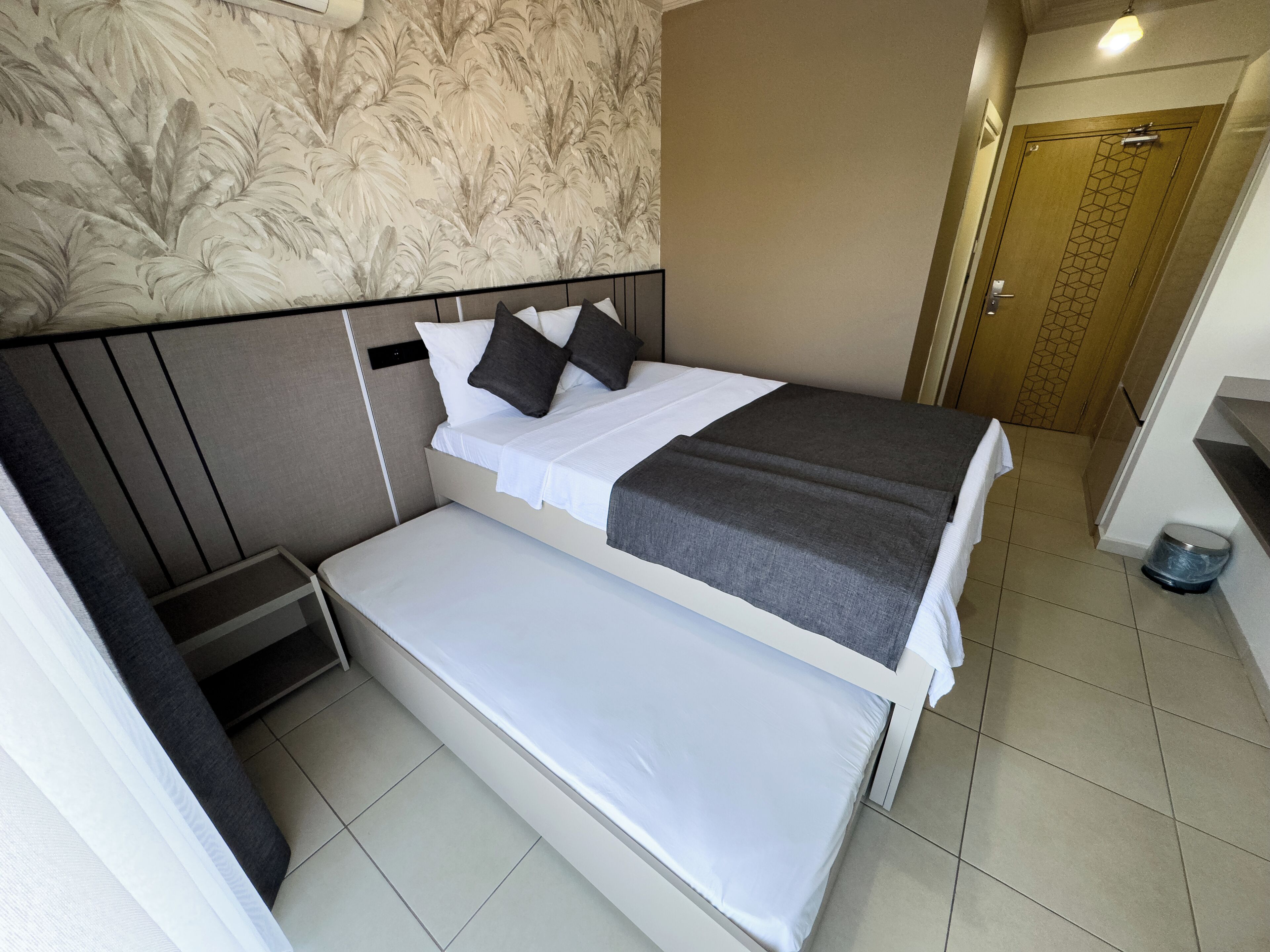 A compact hotel room with a bed, a small bench, a nightstand, and a door.