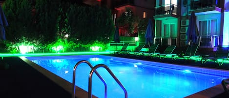 A swimming pool with a metal ladder, surrounded by lounge chairs and umbrellas, under nighttime lighting.