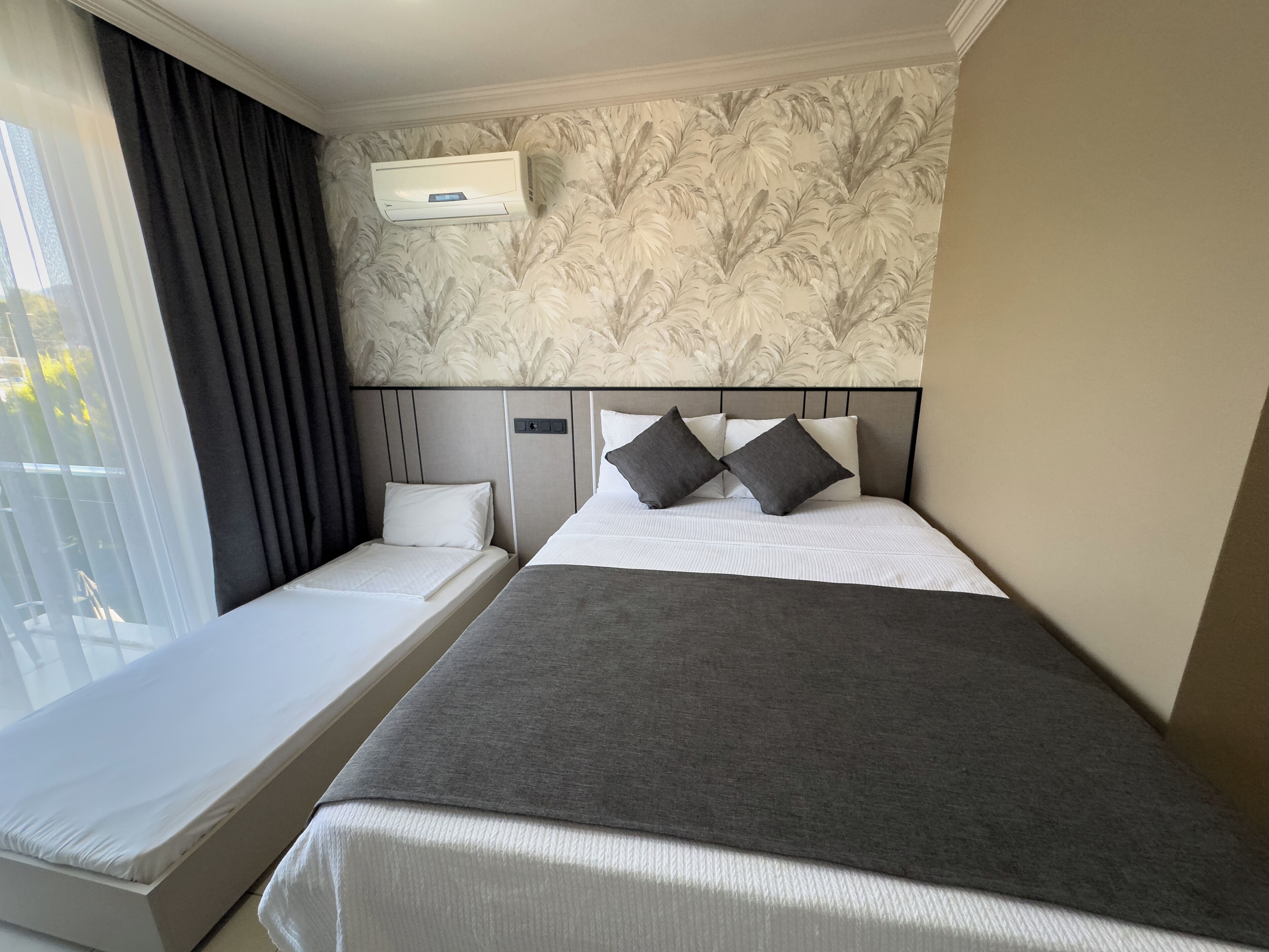 A hotel room with two beds, a large window with curtains, a wall-mounted air conditioner, and a floral wallpaper design.