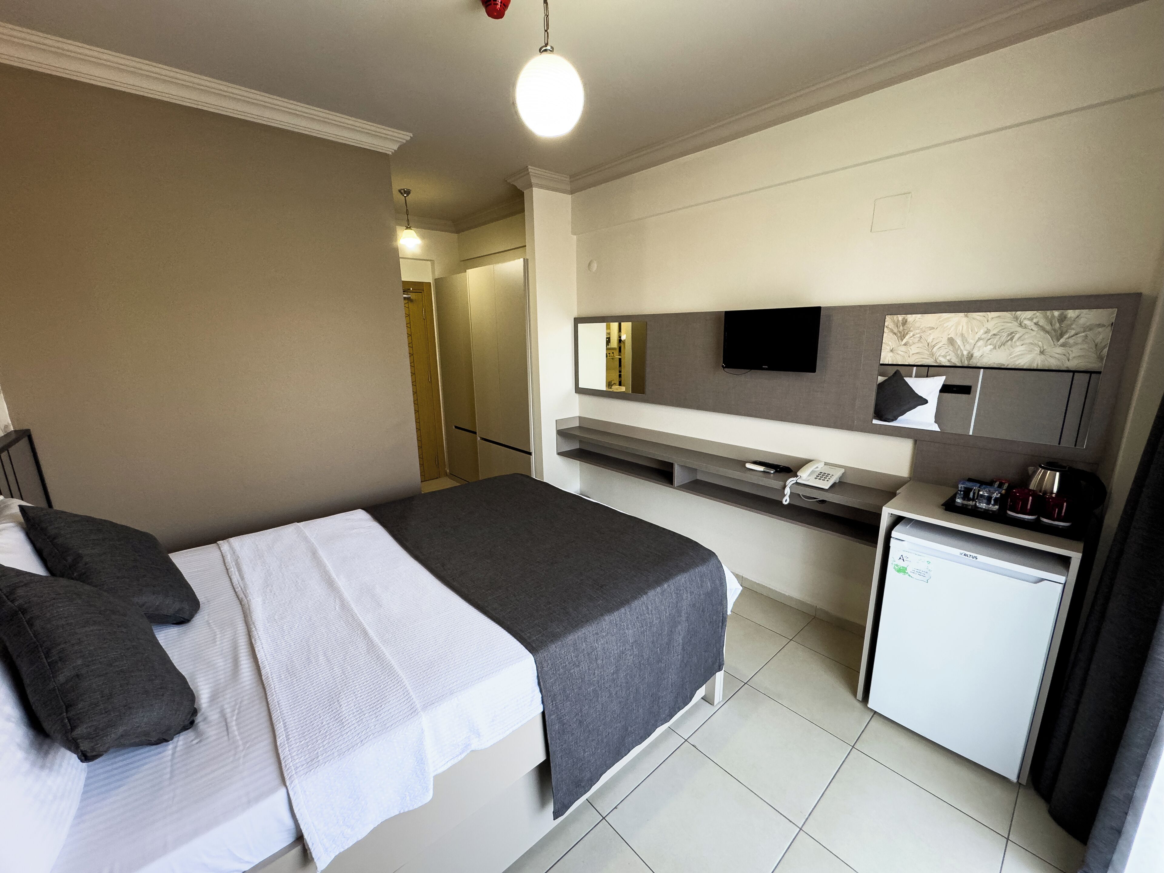 A modern hotel room with a bed, bedside table, a small desk with a telephone, a mirror, and a TV.