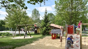 A playground with various play structures, including a swing set and climbing frames, surrounded by trees and a sandy area. - Camping Le Petit Lion - Lodges Bois (Vendoire)