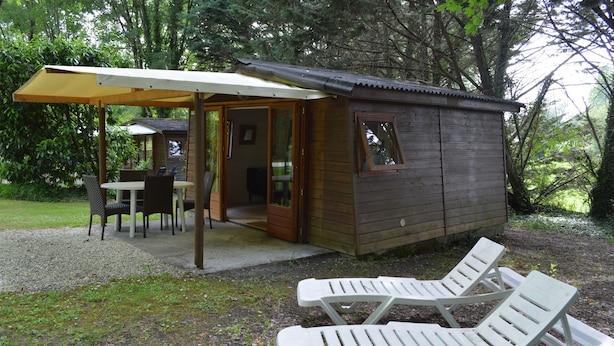 A small wooden cabin with a covered patio and a dining area. There are two white lounge chairs outside. - Camping Le Petit Lion - Lodges Bois (Vendoire)