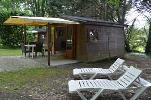 Una pequeña cabaña de madera con un porche cubierto y una zona de comedor. Hay dos sillones blancos afuera.