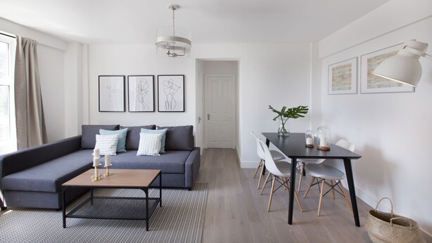 A modern living room with a grey sofa, a coffee table, and a dining area with a table and chairs. There are framed artworks on the wall and a chandelier hanging above the sofa. - Park West by Allô Housing (London)