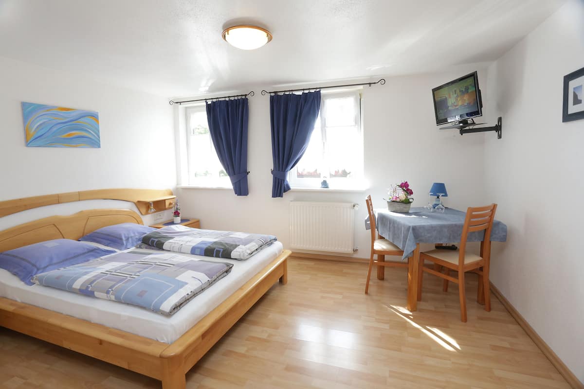 Ein Schlafzimmer mit Bett, Nachttisch, einem Fenster mit Vorhängen, einem kleinen Tisch mit Stühlen und einem an der Wand befestigten Fernseher.