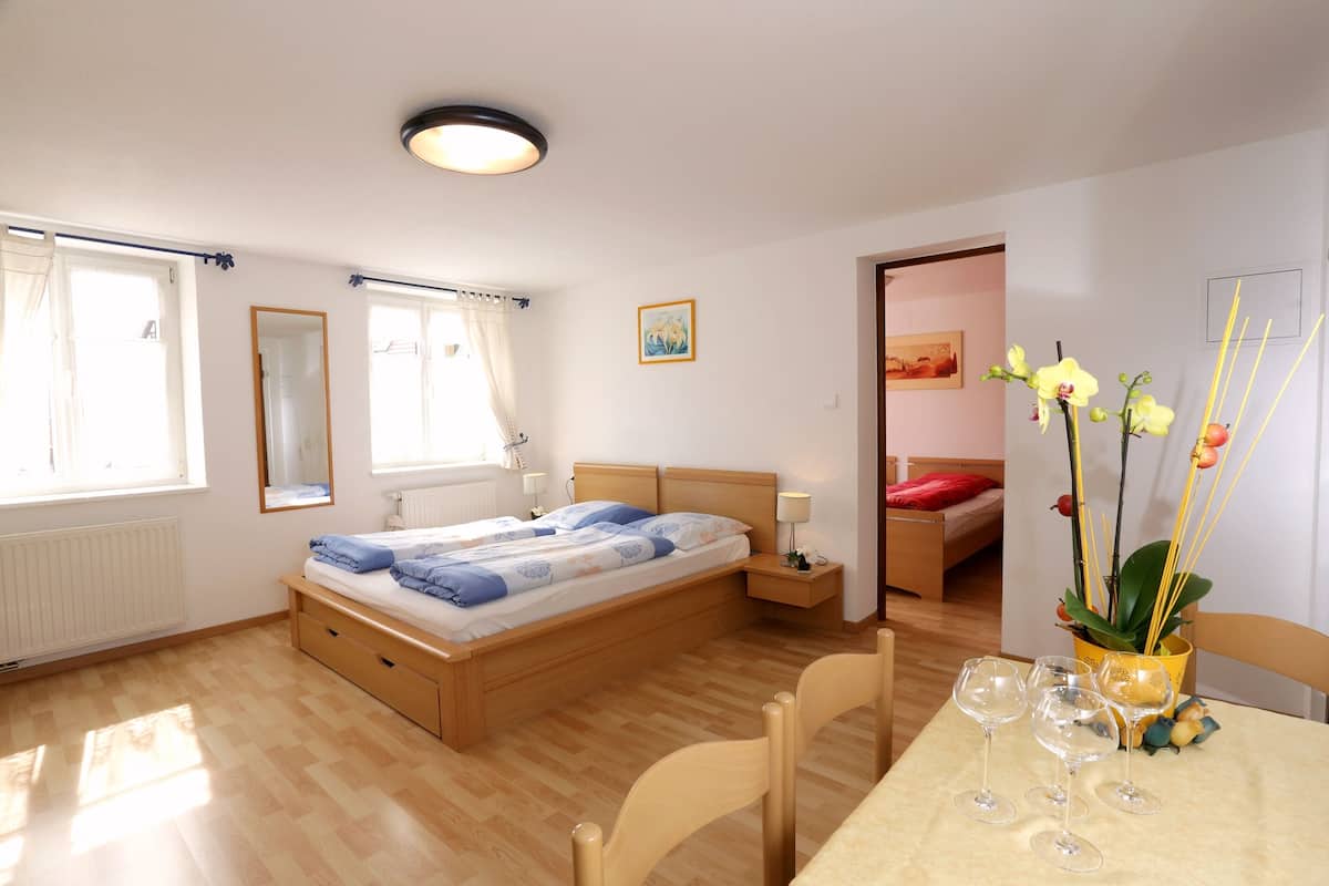 Ein Schlafzimmer mit einem hölzernen Bett, Nachttischen, einem Fenster mit Vorhängen, einem kleinen Tisch mit Weingläsern und einer Tür zu einem weiteren Raum.