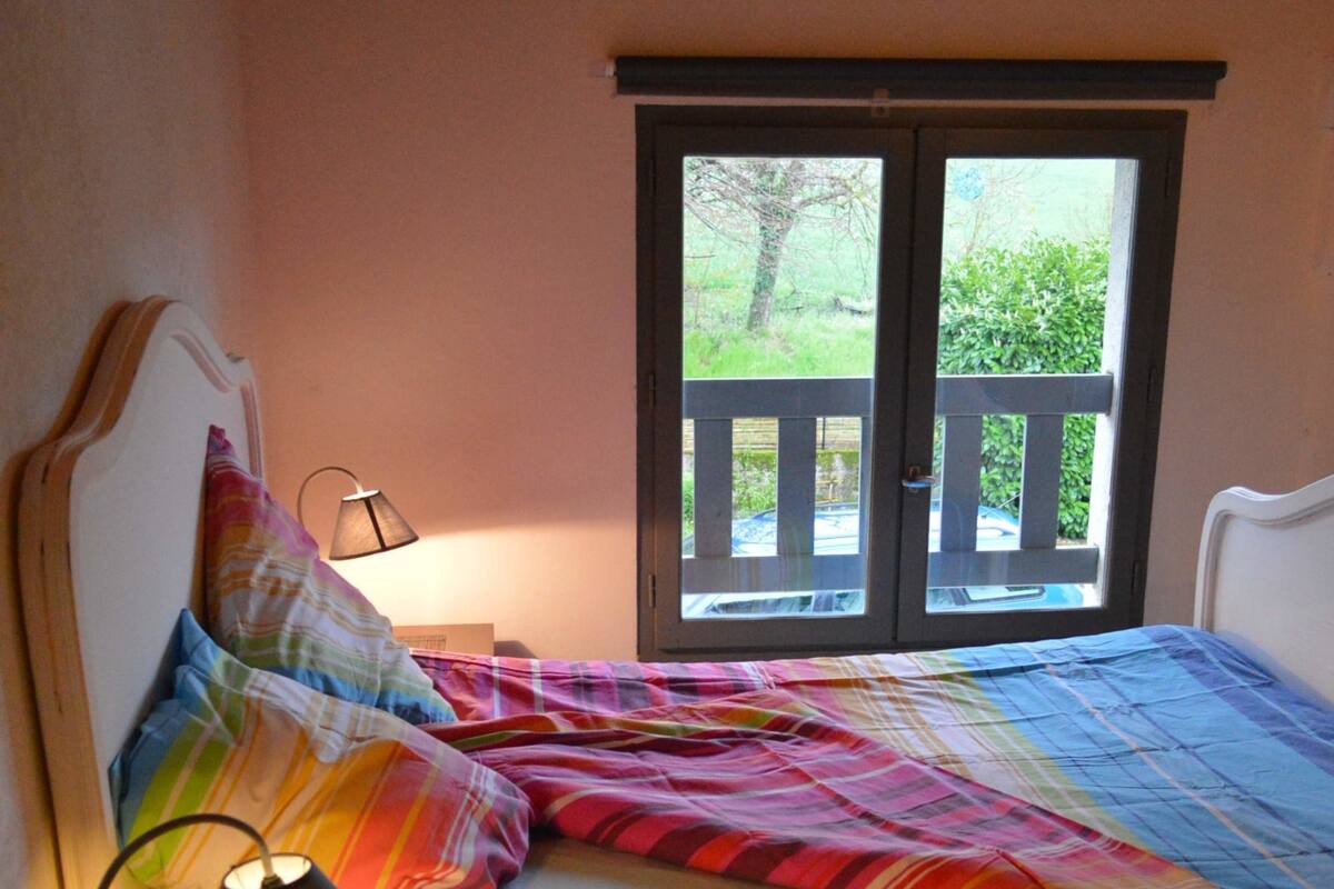 Een slaapkamer met een bed, nachtkastje met een lamp, een boek en een raam met uitzicht op groen.